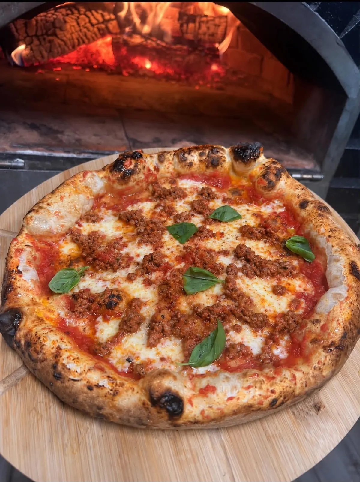 Une pizza à la viande hachée et au basilic, devant les flammes du four à bois