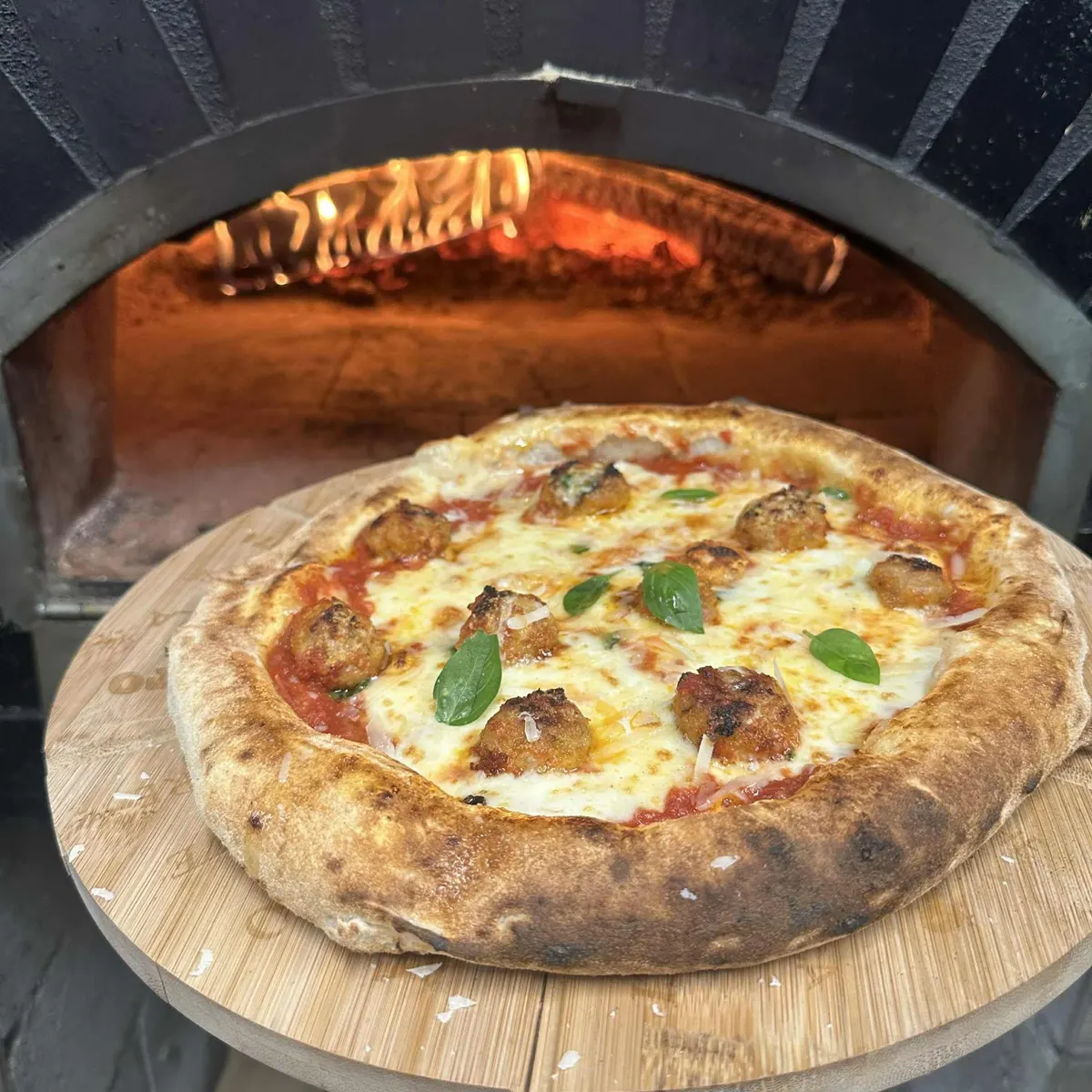 Une pizza aux boulettes de viande et au basilic, sur la pelle, devant le four à bois allumé